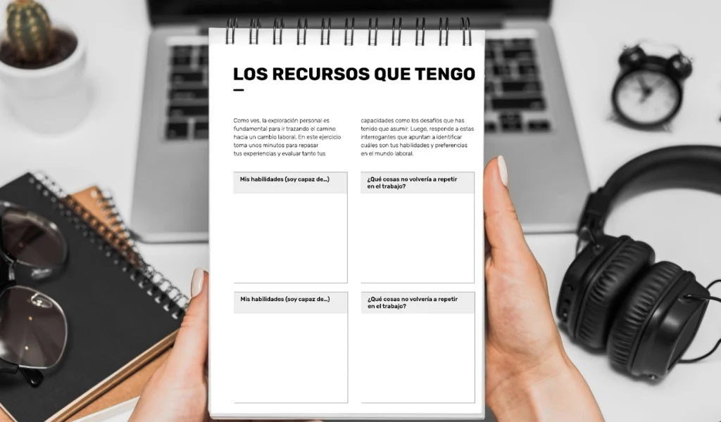 A woman holds the printed version of the planner, in notebook format. In the background, a laptop, a watch, headphones, glasses, notebooks, and a small cactus are visible.