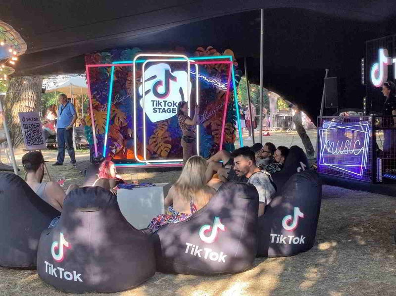 TikTok corner of the festival showing several seats and people sitting and talking. In the background, a small stage with neon lights.